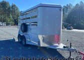 White horse trailer parked in a gravel lot, showing side panels and spare tire with diamond-plate front. A blue sky and pine trees in the background.
