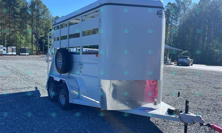 White horse trailer parked in a gravel lot, showing side panels and spare tire with diamond-plate front. A blue sky and pine trees in the background.