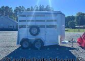 White horse trailer parked on a gravel lot with a spare tire mounted on its rear, blue sky and trees in the background, watermark CarolinaTrailersales.com.