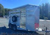 White livestock trailer with open side gate and spare tire mounted on the exterior, parked on a gravel lot with trees in the background.