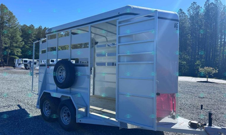 White livestock trailer with open side gate and spare tire mounted on the exterior, parked on a gravel lot with trees in the background.