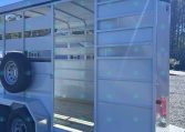 Open livestock trailer with side gate open, showing interior and spare tire mounted on the exterior, under a clear blue sky.
