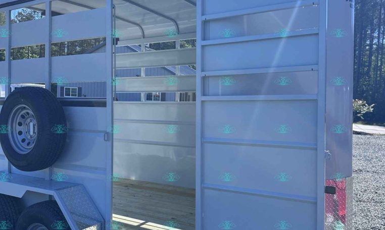 Open livestock trailer with side gate open, showing interior and spare tire mounted on the exterior, under a clear blue sky.