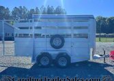 White livestock trailer with a side-mounted spare tire on a gravel lot, trees and blue sky in the background.