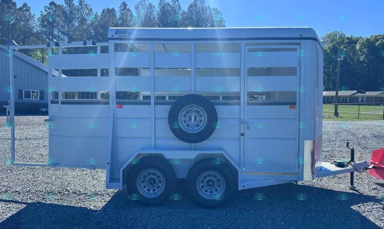 White livestock trailer with a side-mounted spare tire on a gravel lot, trees and blue sky in the background.