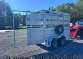 White livestock trailer with ramp down on a gravel lot, dual axles, and a red tractor visible to the right; trees and blue sky in background.