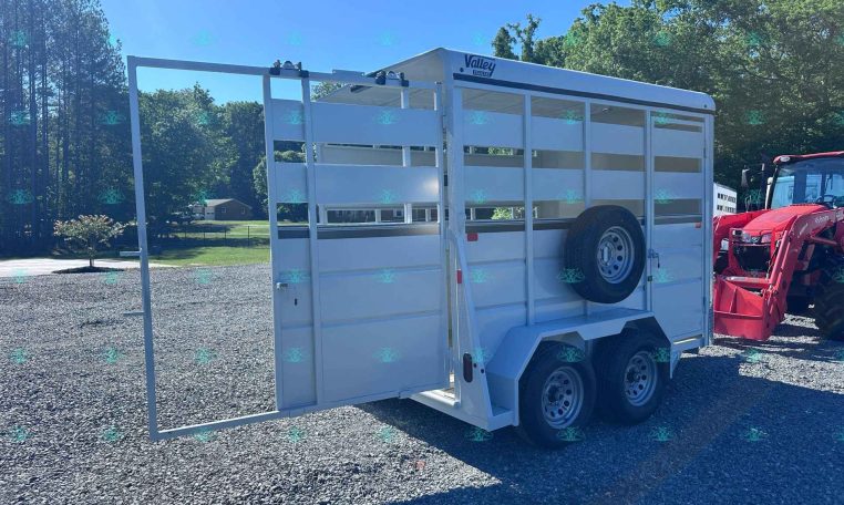 White livestock trailer with ramp down on a gravel lot, dual axles, and a red tractor visible to the right; trees and blue sky in background.