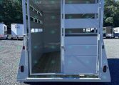 Open horse trailer showing interior stalls with divider rails and a wooden floor, white exterior, door open at the rear, with other trailers on a gravel lot under a clear blue sky