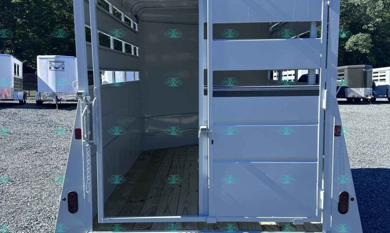 Open horse trailer showing interior stalls with divider rails and a wooden floor, white exterior, door open at the rear, with other trailers on a gravel lot under a clear blue sky