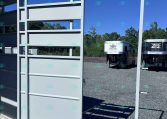 White metal trailer frame/gate on a gravel lot, with horse trailers visible in the background under a blue sky.