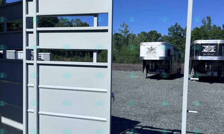 White metal trailer frame/gate on a gravel lot, with horse trailers visible in the background under a blue sky.