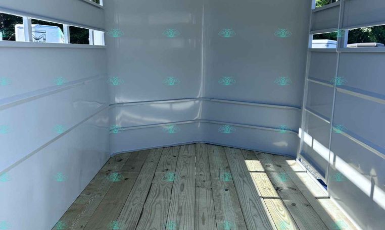 Empty light gray trailer interior with wooden deck and side rails, sunlit from outside.