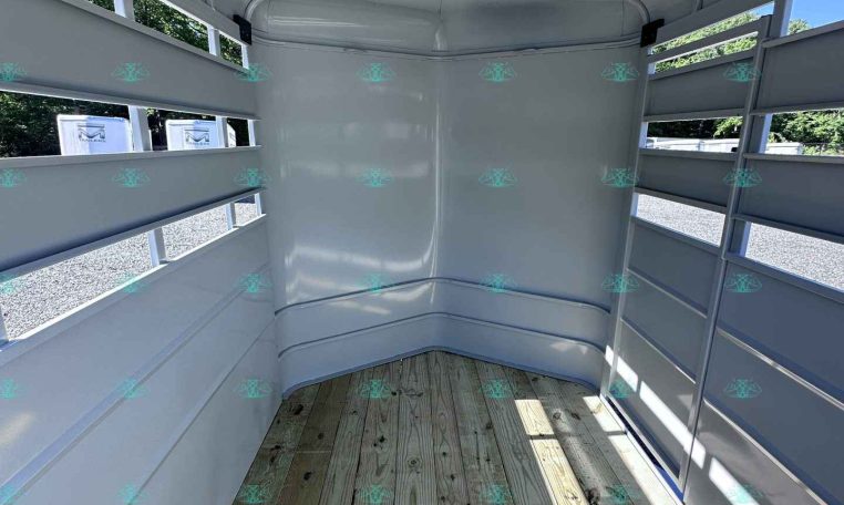 Interior of an empty white horse trailer with wooden floor and side slat windows, sun shining inside