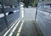Interior view of a light gray horse trailer with back doors open to a gravel lot and trees in the background.