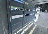 Interior of a light gray horse trailer with a wood plank floor and lowered ramp, open side panels showing other trailers in a gravel lot outside