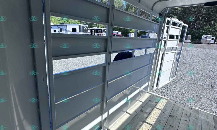 Interior of a light gray horse trailer with a wood plank floor and lowered ramp, open side panels showing other trailers in a gravel lot outside