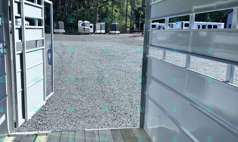 View from inside a white trailer looking out onto a gravel lot with several parked trailers and a wooded backdrop.