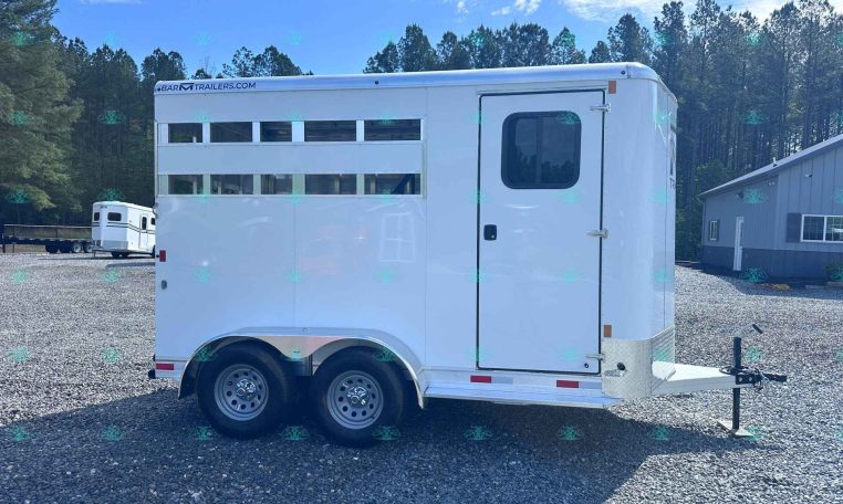 White horse trailer with a side door and sliding windows, parked on a gravel lot with trees in the background and a building nearby. Website watermark CarolinaTrailersales at the bottom.