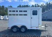 White two‑axle horse trailer with side windows, parked on a gravel lot with trees and a blue sky in the background.