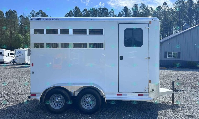 White two‑axle horse trailer with side windows, parked on a gravel lot with trees and a blue sky in the background.