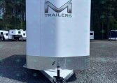 Front view of a white enclosed trailer with a large chrome 'M' logo and the word 'TRAILERS', parked on a gravel lot with trees in the background, hitch visible at the front and diamond-plate trim at the bottom.