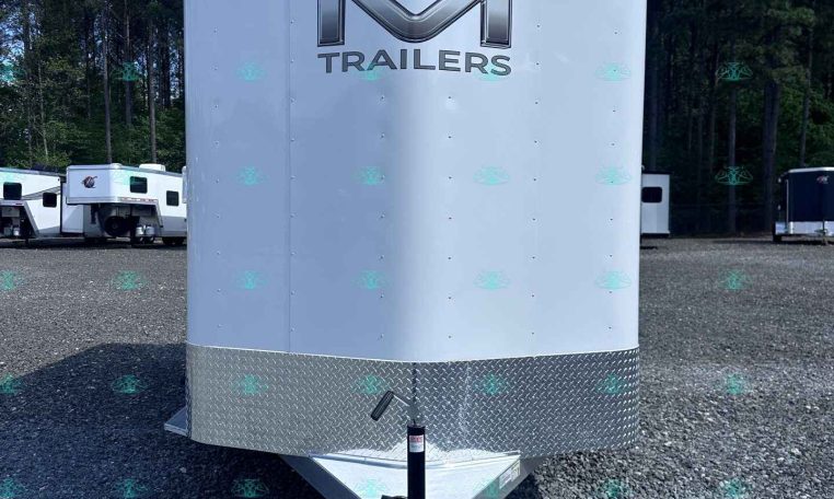 Front view of a white enclosed trailer with a large chrome 'M' logo and the word 'TRAILERS', parked on a gravel lot with trees in the background, hitch visible at the front and diamond-plate trim at the bottom.