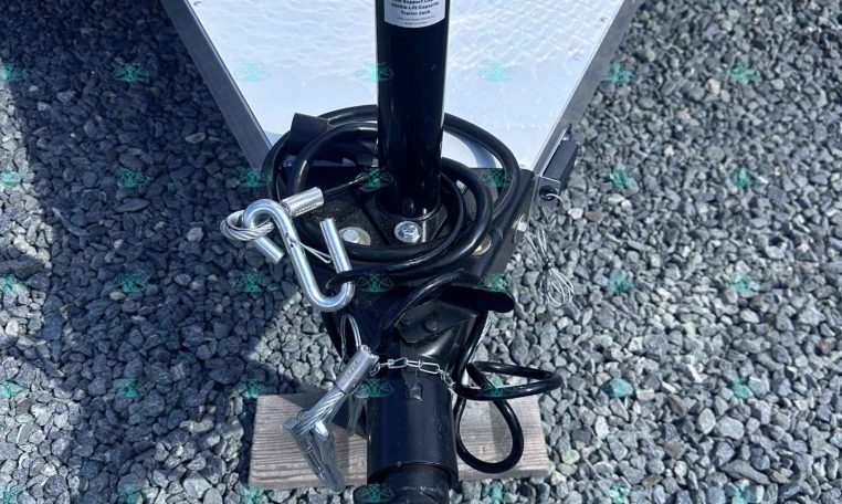 Close-up of a black trailer jack and safety chains attached to a trailer on a gravel surface, with a diamond-plate trailer wall in the background.