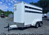 White livestock trailer with two-level ventilation slats and a diamond-plate front, on a gravel lot with other trailers and trees in the background.