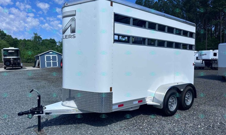 White livestock trailer with two-level ventilation slats and a diamond-plate front, on a gravel lot with other trailers and trees in the background.
