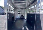 Empty aluminum trailer interior with rear ramp open, bright daylight, side windows, a mounted rack with two black boots, and a spare wheel at the far end inside the trailer.