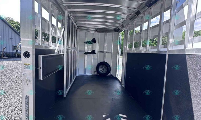 Empty aluminum trailer interior with rear ramp open, bright daylight, side windows, a mounted rack with two black boots, and a spare wheel at the far end inside the trailer.