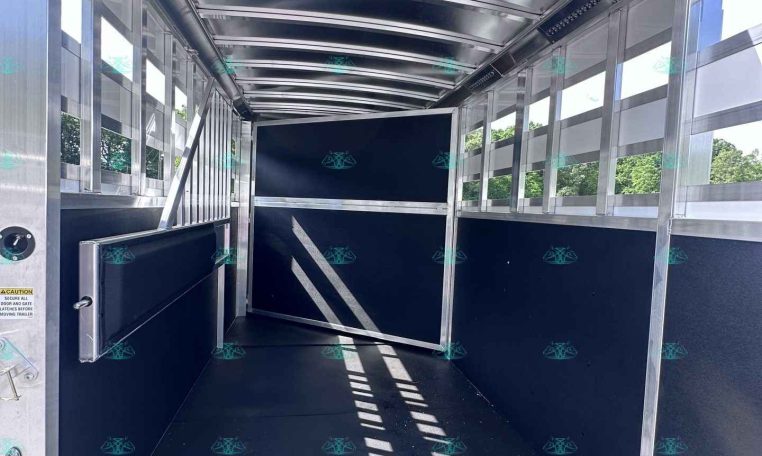 Interior of an empty aluminum trailer with open rear doors, black floor, and metal framing; blue sky visible outside.