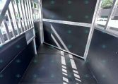 Interior of an empty aluminum trailer with black plywood walls and vertical slats on the left; sunlight creates stripes on the floor.
