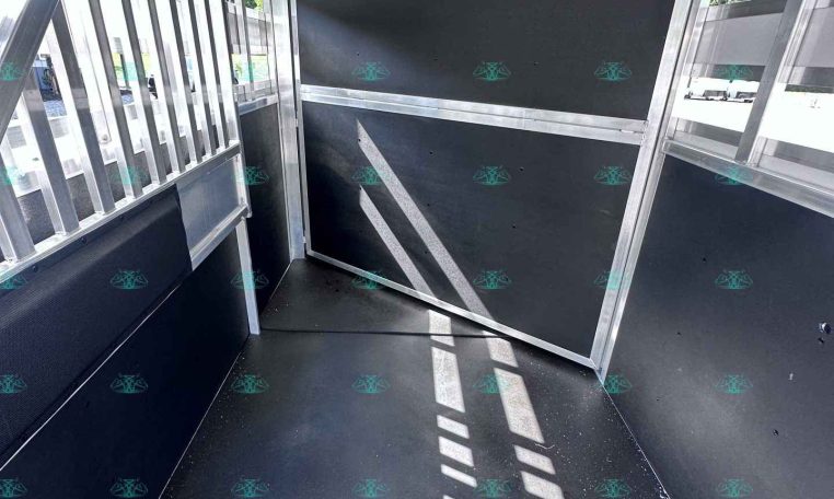 Interior of an empty aluminum trailer with black plywood walls and vertical slats on the left; sunlight creates stripes on the floor.
