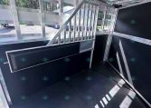Interior of a silver aluminum trailer with exposed framing, black walls, and a metal stair/ramp leading to a higher deck; sunlit windows along one side.