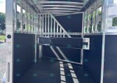 Interior of a silver aluminum horse trailer with a divider and gated stalls, viewed from the open rear doorway in daylight.
