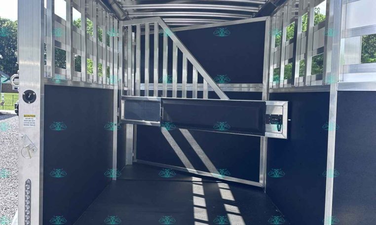 Interior of a silver aluminum horse trailer with a divider and gated stalls, viewed from the open rear doorway in daylight.