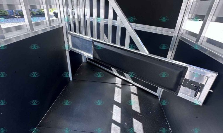 Interior of a metal trailer showing a vertical-bar divider and slanted support beam, black wall panels, and rubberized floor. Light comes from side windows; watermark CarolinaTrailerSales.com visible at bottom.