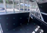 Interior of a metallic trailer showing aluminum framing, a doorway with stairs, and a black interior wall on the right side. The area is well-lit with natural light outside.