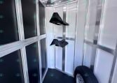 Interior of a metal-framed trailer with overhead hooks along the left wall and a spare tire mounted inside on the right, showing aluminum framing and black wall panels for cargo.