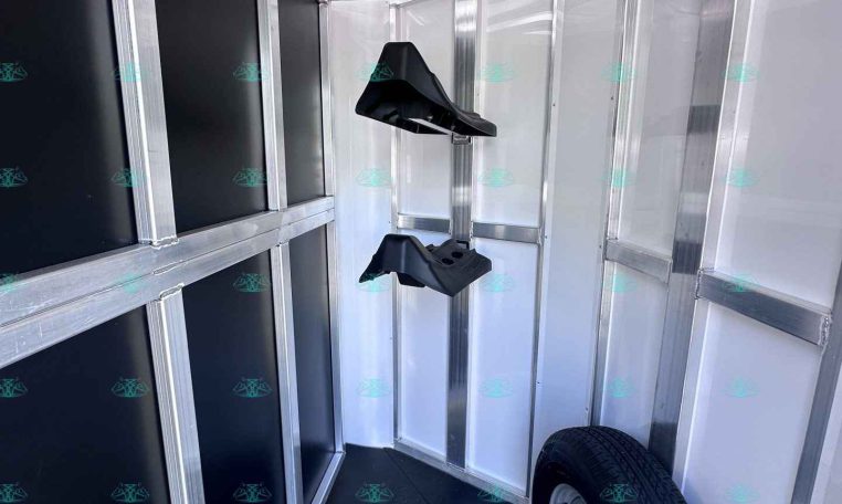 Interior of a metal-framed trailer with overhead hooks along the left wall and a spare tire mounted inside on the right, showing aluminum framing and black wall panels for cargo.