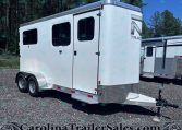 White tandem-axle horse trailer with two side windows on a gravel lot, front hitch visible under clear blue sky.