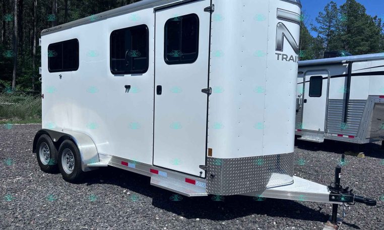 White tandem-axle horse trailer with two side windows on a gravel lot, front hitch visible under clear blue sky.