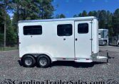 White enclosed trailer with two dark-tinted windows and dual axles parked on a gravel lot, trees in the background.