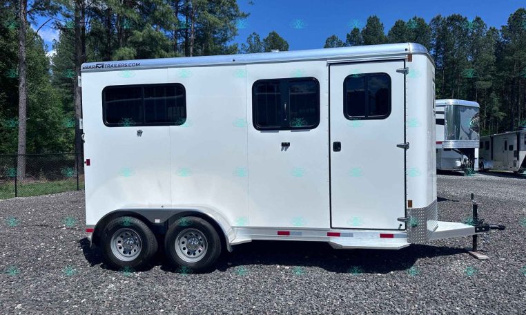 White enclosed trailer with two dark-tinted windows and dual axles parked on a gravel lot, trees in the background.
