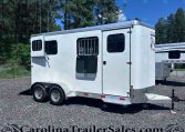 White two-axle horse trailer with barred windows, parked on a gravel lot with a forested backdrop of pine trees.