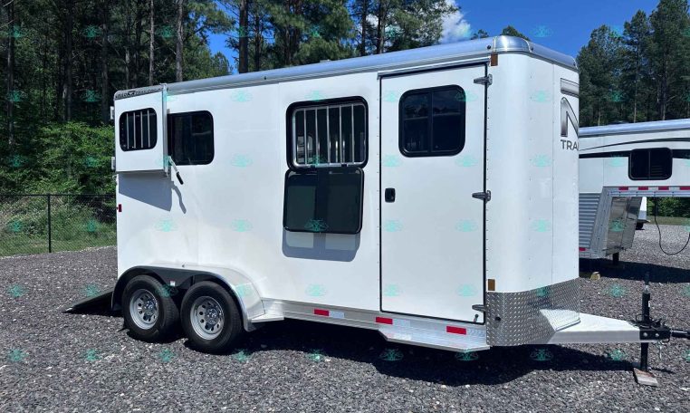 White two-axle horse trailer with barred windows, parked on a gravel lot with a forested backdrop of pine trees.
