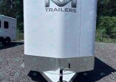 Front view of a white enclosed trailer with a large chrome 'M' emblem and the word 'TRAILERS' on the front, parked on a gravel lot with another trailer in the background.