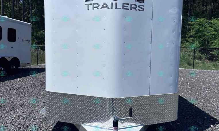 Front view of a white enclosed trailer with a large chrome 'M' emblem and the word 'TRAILERS' on the front, parked on a gravel lot with another trailer in the background.