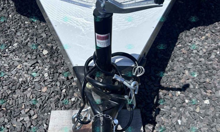 Close-up of a utility trailer hitch showing the jack, coupler, and safety chains on a diamond-plate trailer tongue, resting on gravel.